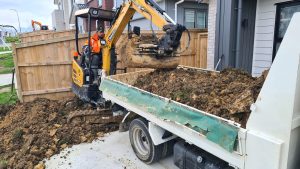 An excavator extracts dirt from a dump truck, set against a backdrop of a concrete driveway and artificial turf installation.
